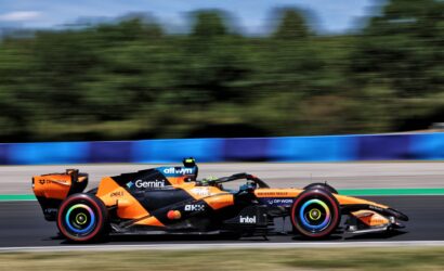 Orange Formula 1 race car speeding along a track, sponsor decals visible along the body with a blurred background for motion.