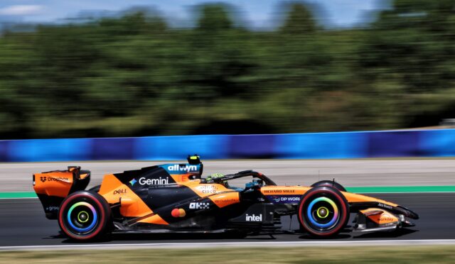 Orange Formula 1 race car speeding along a track, sponsor decals visible along the body with a blurred background for motion.