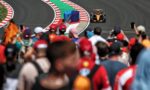 Orange Formula 1 car speeds along a curving track with red-white barriers and a green runoff, blurred crowd in the foreground.