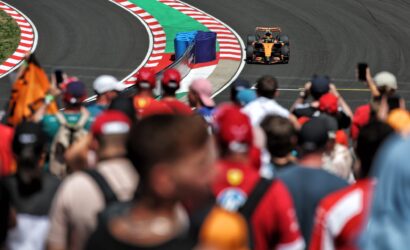 Orange Formula 1 car speeds along a curving track with red-white barriers and a green runoff, blurred crowd in the foreground.