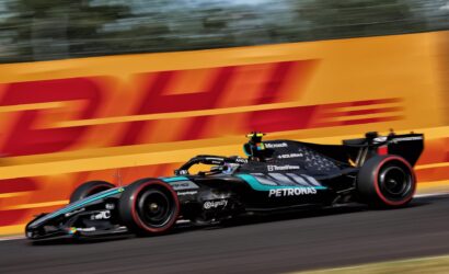 Two Formula 1 cars race at full tilt on a track beside a bright orange DHL barrier, motion blur emphasizing speed.