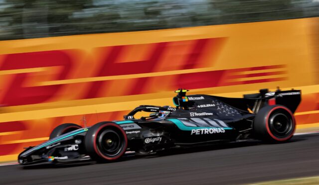 Two Formula 1 cars race at full tilt on a track beside a bright orange DHL barrier, motion blur emphasizing speed.