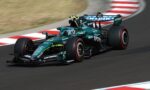 Teal Formula 1 car speeds around a turn on a race track with red-and-white curbs in the background.