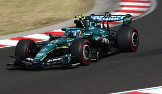 Teal Formula 1 car speeds around a turn on a race track with red-and-white curbs in the background.