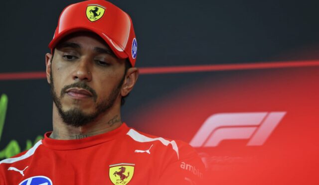 Man in a red Ferrari cap and shirt at a press event, looking slightly downward with a dark backdrop behind and a red glow.