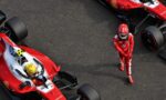 Overhead view of two red Formula 1 race cars on the tarmac with a driver in a red racing suit standing nearby as a pit area scene unfolds.