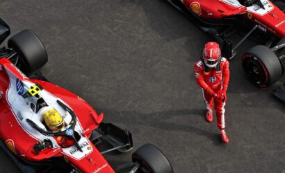 Overhead view of two red Formula 1 race cars on the tarmac with a driver in a red racing suit standing nearby as a pit area scene unfolds.
