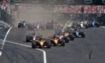 Wide shot of a Formula 1 starting grid racing off the line, cars kicking up dust as they accelerate under an AWS banner.