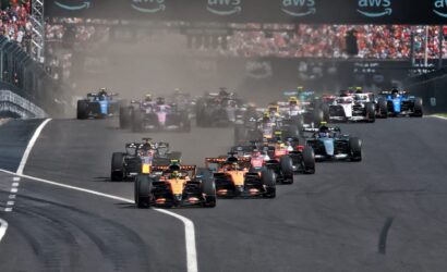 Wide shot of a Formula 1 starting grid racing off the line, cars kicking up dust as they accelerate under an AWS banner.
