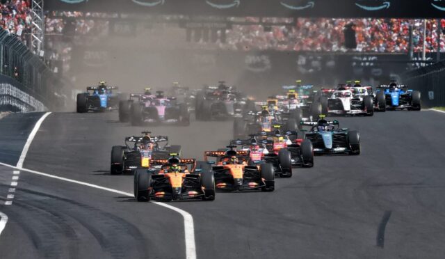 Wide shot of a Formula 1 starting grid racing off the line, cars kicking up dust as they accelerate under an AWS banner.