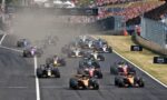 Formula 1 race start: a dense grid of colorful cars on a straight, dust rising behind them, with packed stands and AWS banners in view.