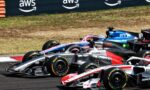 Multiple Formula 1 cars race in close formation on a sunlit track, with sponsor logos on silver, pink, and blue liveries and a dark barrier in the background.