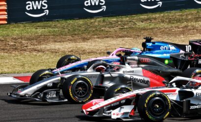 Multiple Formula 1 cars race in close formation on a sunlit track, with sponsor logos on silver, pink, and blue liveries and a dark barrier in the background.