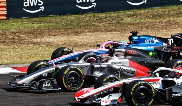 Multiple Formula 1 cars race in close formation on a sunlit track, with sponsor logos on silver, pink, and blue liveries and a dark barrier in the background.