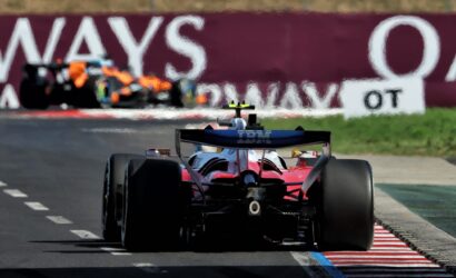 Red Formula 1 car seen from behind on a race track, IBM branding on the rear wing, approaching striped red‑and‑white curbs with a blurred pit wall in the background