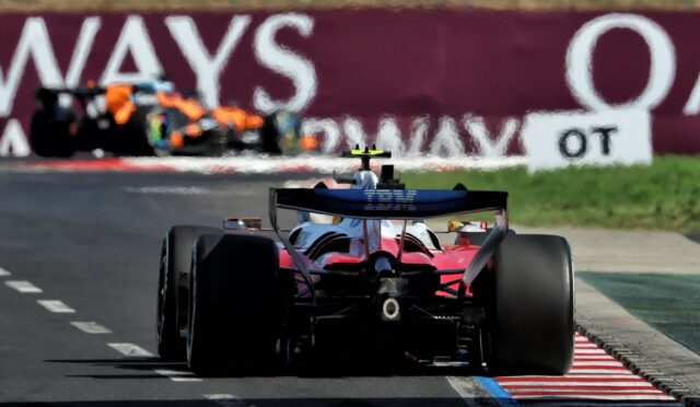 Red Formula 1 car seen from behind on a race track, IBM branding on the rear wing, approaching striped red‑and‑white curbs with a blurred pit wall in the background