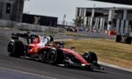 Red Formula 1 race car (#44) rounding a left-hand corner on a race track, with barriers and buildings in the background, speed blur evident.