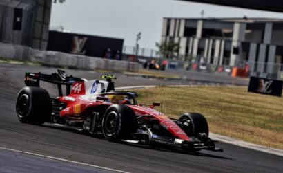 Red Formula 1 race car (#44) rounding a left-hand corner on a race track, with barriers and buildings in the background, speed blur evident.