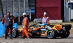 Orange Formula race car at the pit area with three crew members in orange overalls and white helmets nearby, next to a chain-link fence.