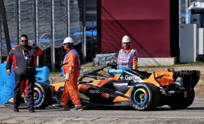 Orange Formula race car at the pit area with three crew members in orange overalls and white helmets nearby, next to a chain-link fence.