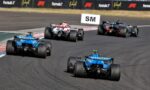 Open-wheel race cars on a track in a tight bend, blue cars following a red-and-white car with sponsor banners visible in the background.