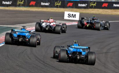 Open-wheel race cars on a track in a tight bend, blue cars following a red-and-white car with sponsor banners visible in the background.