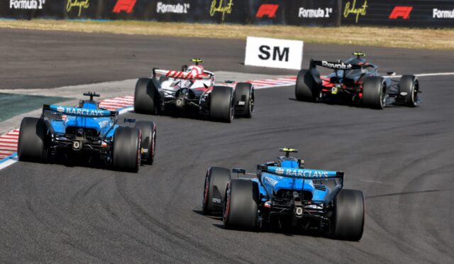 Open-wheel race cars on a track in a tight bend, blue cars following a red-and-white car with sponsor banners visible in the background.