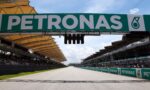 Large PETRONAS sign spans the race track above, with grandstands and spectators on either side."Can be clearer: 'Large PETRONAS sign spans the track above a racing venue'