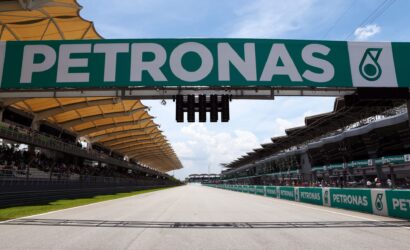 Large PETRONAS sign spans the race track above, with grandstands and spectators on either side."Can be clearer: 'Large PETRONAS sign spans the track above a racing venue'