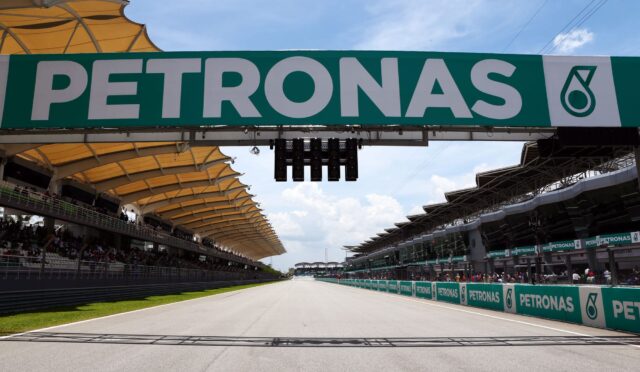 Large PETRONAS sign spans the race track above, with grandstands and spectators on either side."Can be clearer: 'Large PETRONAS sign spans the track above a racing venue'