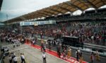 Crowd-filled grandstand at a Formula-style race track with a busy pit area and banners overhead (Petronas Primax).