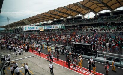 Crowd-filled grandstand at a Formula-style race track with a busy pit area and banners overhead (Petronas Primax).