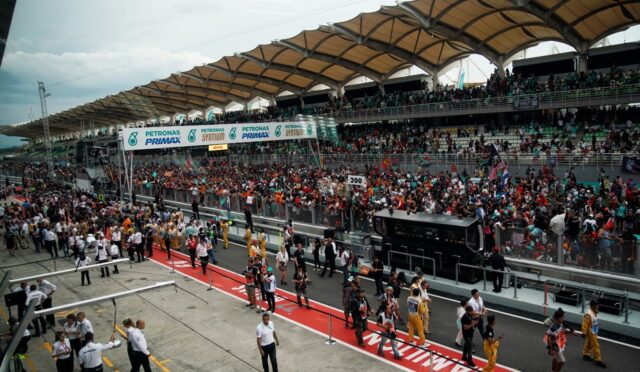 Crowd-filled grandstand at a Formula-style race track with a busy pit area and banners overhead (Petronas Primax).