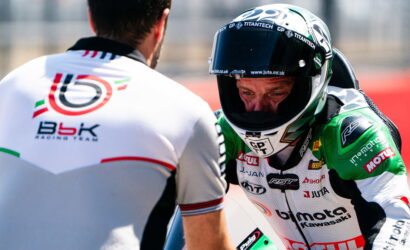 Moto racer in a white-and-green racing suit and full-face helmet talks with a crew member on a racetrack, back of the crewman visible in foreground.