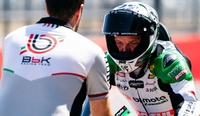 Moto racer in a white-and-green racing suit and full-face helmet talks with a crew member on a racetrack, back of the crewman visible in foreground.