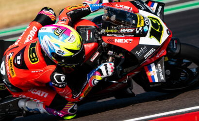 MotoGP rider in a red suit leans into a sharp track corner on a red race bike, colorful helmet visible.