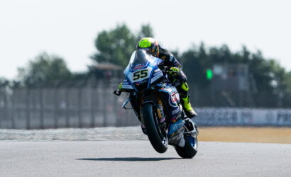 Motorcycle racer in a neon helmet leaning into a turn on a blue race bike number 55, front wheel lifted on a sunlit track.