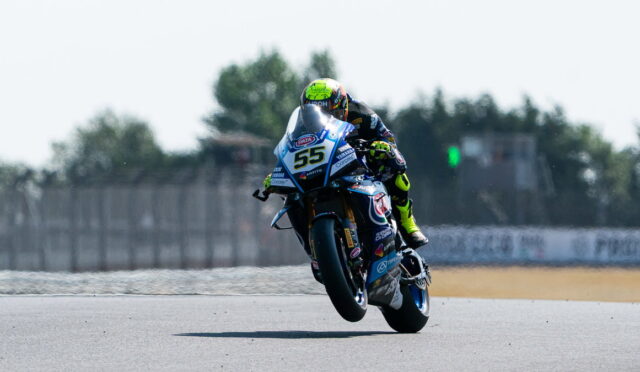 Motorcycle racer in a neon helmet leaning into a turn on a blue race bike number 55, front wheel lifted on a sunlit track.