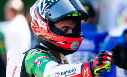 Motorcycle racer wearing a bright red helmet and green racing suit, extending a fist bump toward another rider.