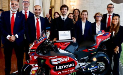 Group of professionally dressed people posing with a red Ducati racing motorcycle, center figure holding a certificate amid sponsor logos (Lenovo, Shell).