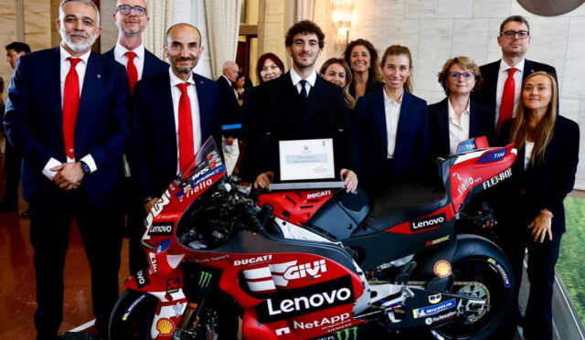 Group of professionally dressed people posing with a red Ducati racing motorcycle, center figure holding a certificate amid sponsor logos (Lenovo, Shell).