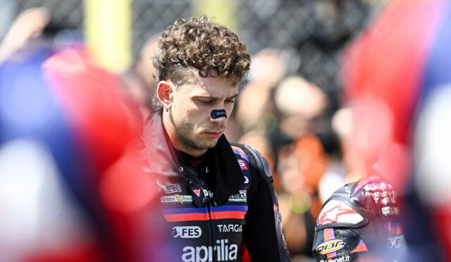 Motorcycle racer with curly hair in a red-and-black suit, looking down as spectators blur in the background.