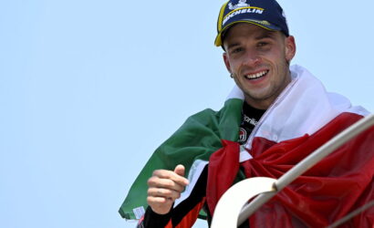 Smiling man wrapped in the Italian flag, wearing a blue Michelin cap, against a clear blue sky.