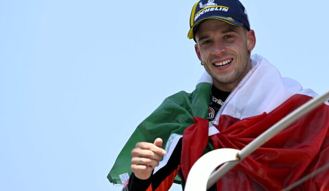 Smiling man wrapped in the Italian flag, wearing a blue Michelin cap, against a clear blue sky.