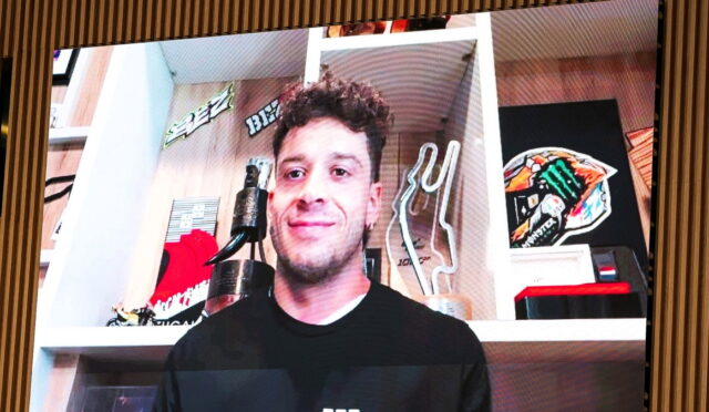 Smiling man in a black shirt standing in a room with shelves, trophies, and decorative items behind him.