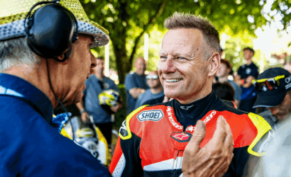 Motorsport rider in a red racing suit smiles while talking with an older man wearing a headset at an outdoor event with spectators in the background.