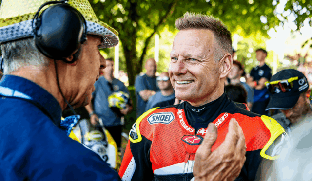 Motorsport rider in a red racing suit smiles while talking with an older man wearing a headset at an outdoor event with spectators in the background.