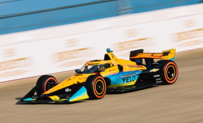 Bright orange and blue open-wheel race car speeds along a race track, sponsors visible on the bodywork.