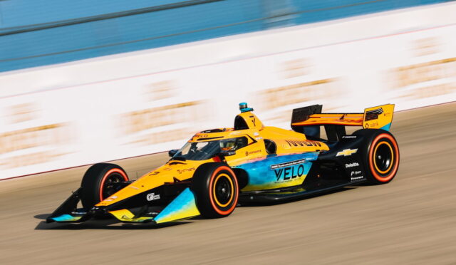 Bright orange and blue open-wheel race car speeds along a race track, sponsors visible on the bodywork.