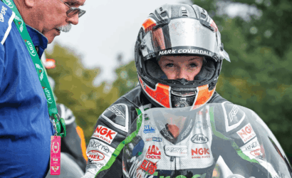 Person in a full racing suit and helmet with sponsor logos stands beside an official in a blue shirt and green lanyard.